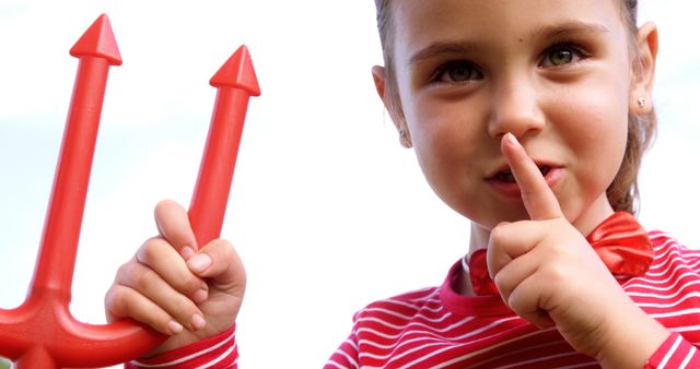 A young Caucasian girl holds up two red toy arrows, one in each hand, and places a finger to her lips in a gesture for silence, with copy space. Her playful expression and the oversized toys suggest a moment of fun and imagination.