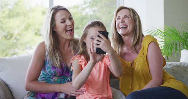 Three females, two adults and a young girl, are enjoying time together as they take a selfie in a bright living room. The scene appears casual with the women laughing and exhibiting positive emotions while the girl captures the moment on her smartphone. Ideal for use in family-related content, lifestyle blogs, social media promotions, or advertisements focusing on bonding and togetherness.