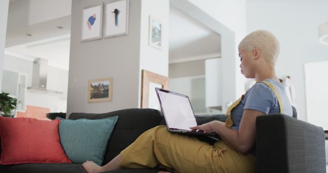 Woman sitting on couch working on laptop in cozy living room. She is wearing casual clothes and is focused on her work. Ideal for use in articles related to work-from-home setups, remote work lifestyle, modern home interiors, and technology use at home.