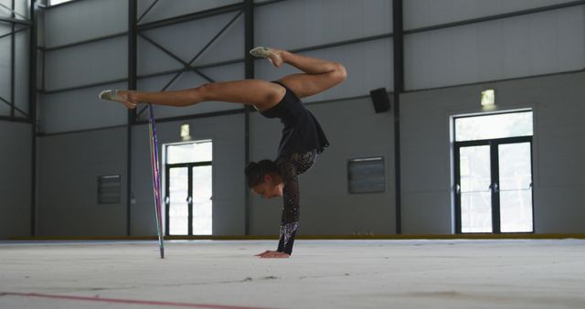 Rhythmic gymnast practicing on mat in gymnasium, performing routine with ribbon. Athlete showcases balance, flexibility, and coordination in professionally equipped facility. Suitable for use in promotional materials for gymnastics events, sportswear advertisements, athletic training programs, and articles on fitness and training disciplines.