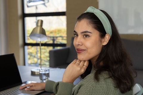 Young woman working remotely at home desk with laptop, wearing green sweater and headband