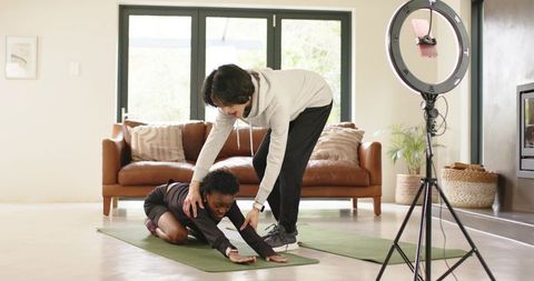 Home fitness coach assisting african american woman stretching on yoga mat with ring light