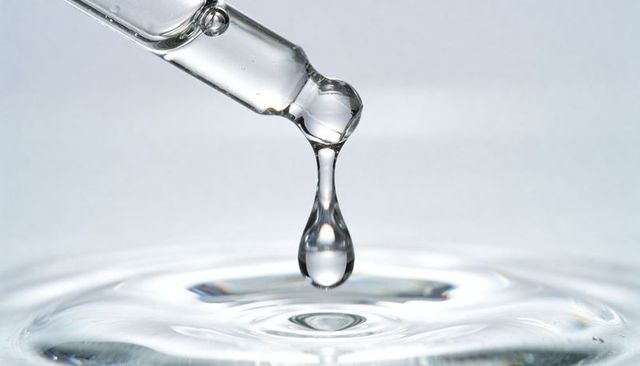 Glass pipette dripping single clear droplet over gentle rippling water surface laboratory