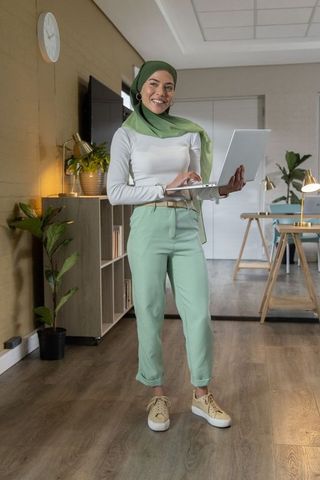 Smiling woman in green hijab standing with laptop in modern home office for remote work