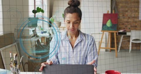 Mature woman working on laptop in art studio with digital ui overlay and easel