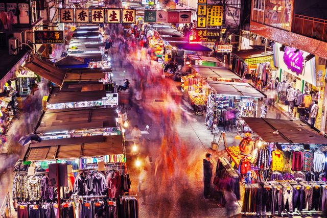 Showcasing a busy night market with numerous colorful stalls and a dynamic crowd. Asian city ambience and street market vibrancy highlighted by blurred motion of people walking. Perfect for travel guides, cultural blogs, and articles on urban nightlife or traditional markets.