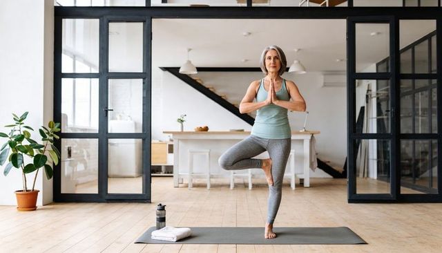 Senior woman practicing tree pose on yoga mat in bright modern open-plan home