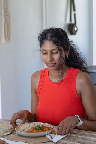 Woman wearing red tank top eating rice and curry at wooden table in minimal home interior
