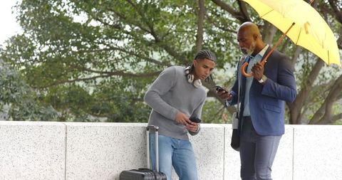 Urban businessman and young traveler checking phones under yellow umbrella by suitcase