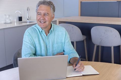 Mature man working from home in modern kitchen smiling while typing and writing notes