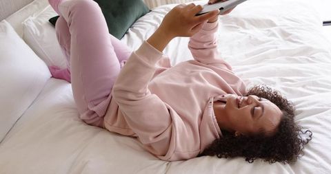 African american woman lying on bed in pink loungewear holding tablet and smiling, cozy leisure