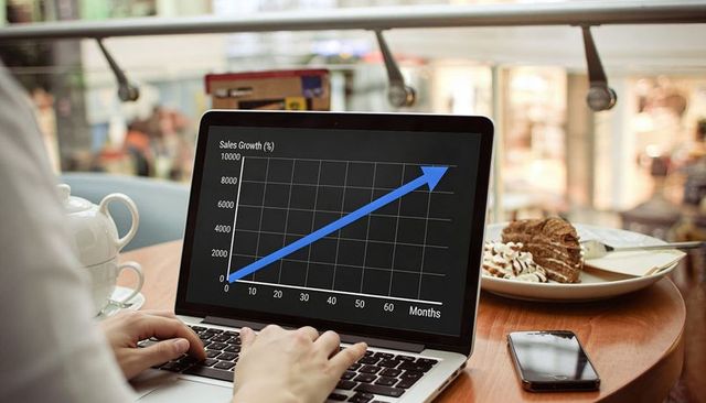 Entrepreneur tracking sales growth on laptop in cafe with smartphone and dessert