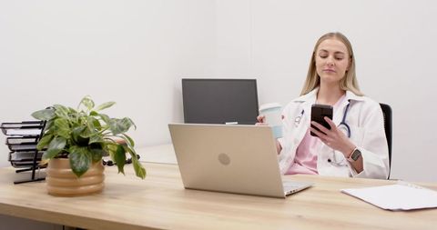 Young female doctor checking smartphone while holding coffee at clinic desk with laptop