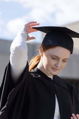 Female graduate adjusting mortarboard cap wearing black gown and white top on sunlit campus