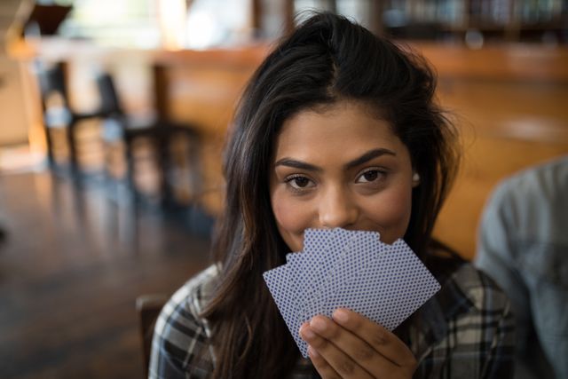 Young woman smiling while holding playing cards in a bar. Ideal for use in content related to leisure activities, social gatherings, gaming, and casual settings. Perfect for illustrating themes of fun, relaxation, and social interaction.