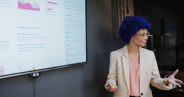 Young professional with blue hair and glasses presenting data on a large wall screen in an office. The screen displays various graphs and statistical reports. Ideal for use in business presentations, career development campaigns, workplace diversity, and modern office environments.