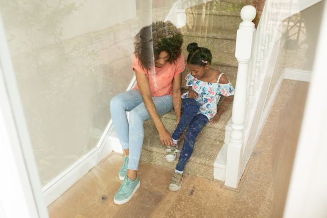 African American woman enjoying her time at home with her daughter, sitting on staircase mother helping daughter putting her shoes on. Family togetherness domestic life.