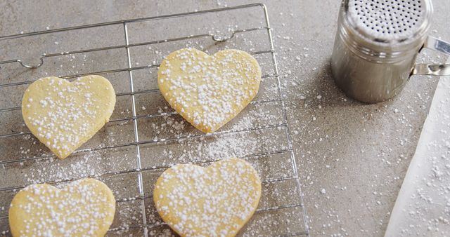 Heart-shaped sugar cookies on a cooling rack scattered with powdered sugar. A powdered sugar sifter nearby suggests fresh baking. Perfect for illustrating Valentine’s Day, home baking, holiday treats, or cooking sessions.