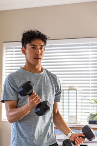 Asian man working out at home lifting hex dumbbells by sunlit window blinds