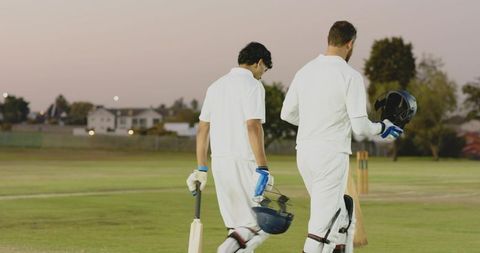 Twilight cricket teammates in white kit walking across outfield carrying bats and helmets