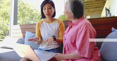 Young Asian woman teaching older adult laptop skills on sunlit patio, family tech help