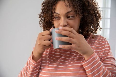 African American Woman Sipping Hot Beverage with Sideways Gaze Near Blinds