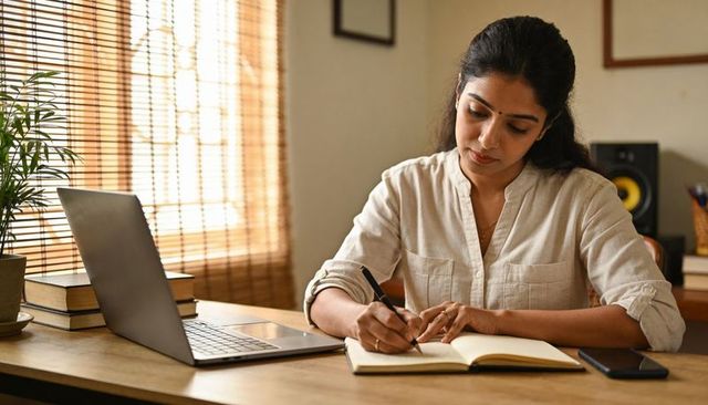 Indian woman writing in notebook at sunlit minimalist wooden desk with laptop and phone