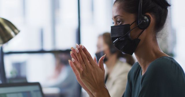 Biracial businesswoman wearing face mask at desk using computer and talking wearing phone headset. independent creative business in a modern office during coronavirus covid 19 pandemic.