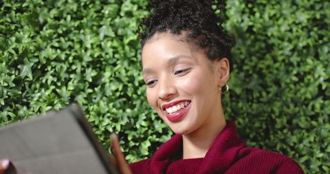 Smiling young woman using tablet against leafy hedge in maroon sweater