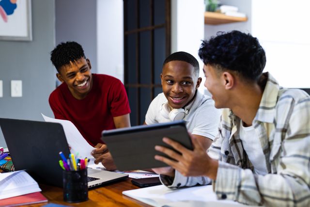 Teenage friends are gathered at home, using tablets and laptops. They are smiling and engaging in conversation, suggesting a collaborative and friendly atmosphere. This image is ideal for illustrating concepts related to friendship, teamwork, modern education, and leisure activities among teenagers. It can be used in advertisements, educational materials, and social media posts promoting youth engagement and technology use.
