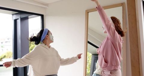 Diverse friends dancing while applying face masks in loungewear by mirror at home