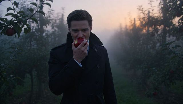 Man biting red apple in misty orchard wearing double-breasted overcoat and tie at dawn
