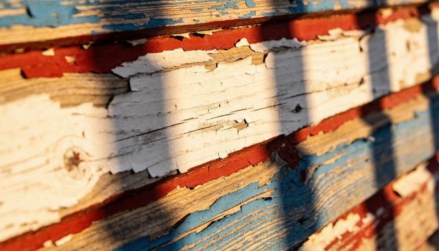 Sunlit peeling red white blue clapboard siding showing weathered wood grain and patina