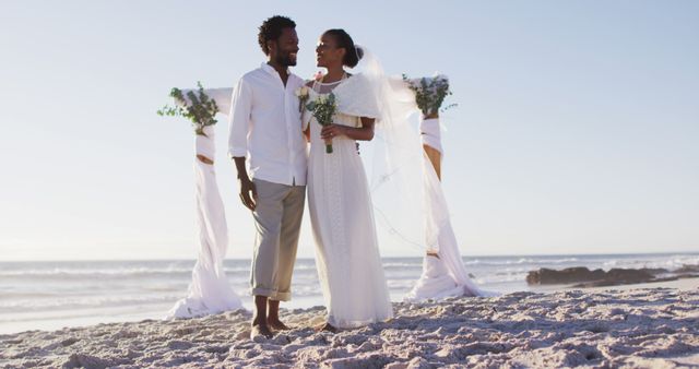 This stock photo depicts a happy Black couple celebrating their wedding on a beach at sunset. They stand together in their casual yet elegant attire, with the bride holding a bouquet and wearing a veil over her white dress. Ideal for articles, blogs, or promotional materials related to beach weddings, destination ceremonies, or romantic celebrations outdoors. The beautiful ocean backdrop underlines the serene and joyful atmosphere of the event.