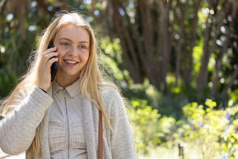 Blonde woman smiling while talking on smartphone outdoors in sunlit green park wearing cardigan