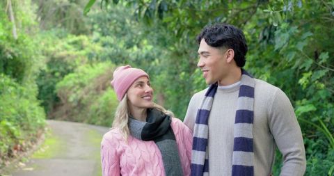 Diverse couple strolling and smiling on woodland trail wearing knit sweaters and scarves