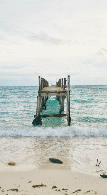 Old wooden pier extending into calm turquoise ocean water. Ideal for concepts related to tranquil beach vacations, seaside relaxation, nature's beauty, and serene landscapes. Suitable for travel promotions, outdoor adventure advertisements, or background within aquatic-themed projects.