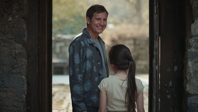 Smiling man and young girl at rustic stone doorway overlooking sunlit cobblestone courtyard