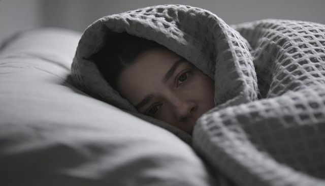 Moody portrait of woman lying under waffle gray blanket, peeking from hooded cover