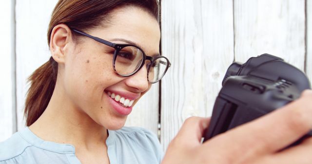 Woman, wearing glasses, smiling while holding and using a DSLR camera. Perfect for showcasing passion for photography, hobbies, and expressing happiness through photography. Suitable for websites, blogs, or advertisements related to photography, eyeglasses, hobbies, lifestyle, or personal stories.