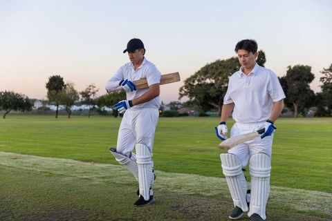 Two cricketers walking across pitch at dusk carrying bats and wearing pads