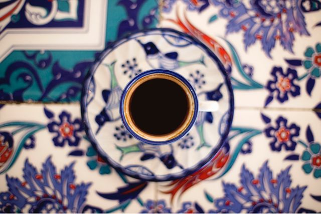Showcasing a top view closeup of Turkish coffee served in an intricately decorated mug placed on a vibrant, patterned table. Highlighting the traditional middle eastern motifs, perfect for themes related to cultural heritage, traditional recipes, coffee culture, and artistic designs. Ideal for bloggers, content creators, or anyone looking to infuse an authentic touch to any coffee-based visual content.