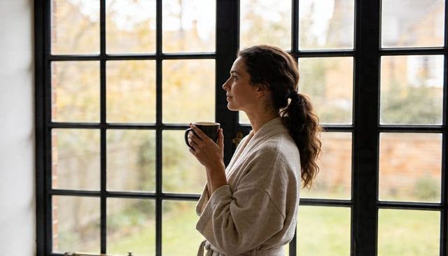 Woman in robe holding coffee by paned window standing contemplative in morning light