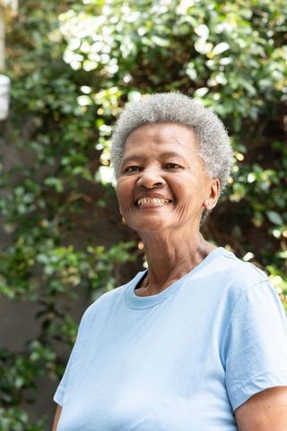 Senior black woman smiling and standing in sunlit garden wearing light blue t-shirt and studs