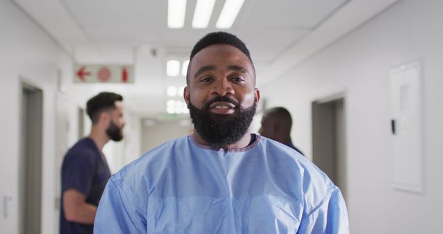 Image portrait of smiling, bearded african american male medical worker in surgical gown in corridor. Hospital, medical and healthcare services.