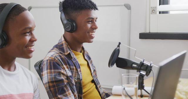 Two teenage boys are engaged in podcasting in a modern studio environment. They are smiling while wearing headphones and speaking into a microphone, suggesting they are enjoying the activity. Ideal for illustrating themes related to youth, technology, modern communication, teamwork, and creativity in media projects. Suitable for blogs, educational content, advertisements, and promotional material focused on teenage engagement, podcasting, or media production.