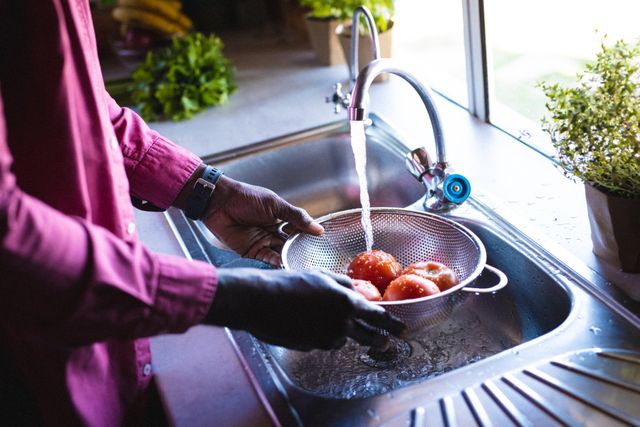 Midsection of african american senior man washing tomatoes in colander under running water in sink. Unaltered, retirement, vacation, solitude, hand, preparation, food, faucet, cleaning concept.