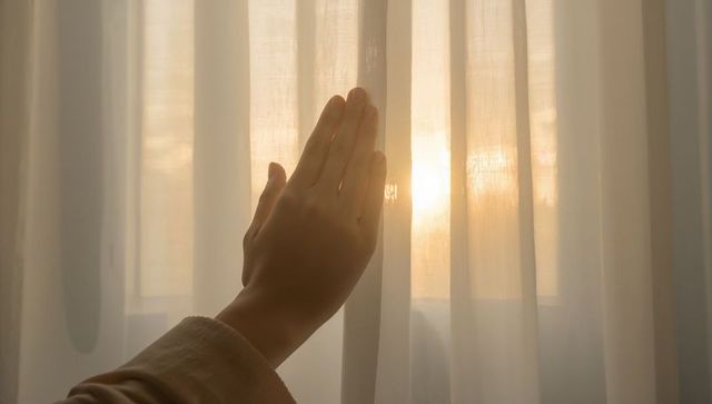 Hand reaching through sheer curtain into warm morning sunlight