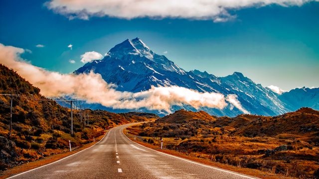 Two-lane highway stretching towards towering snow-capped mountain with blue and cloudy sky. Ideal for content related to travel, adventure, road trips, nature exploration, outdoor activities, and tourism. Perfect for inspirational and motivational themes or travel magazines.