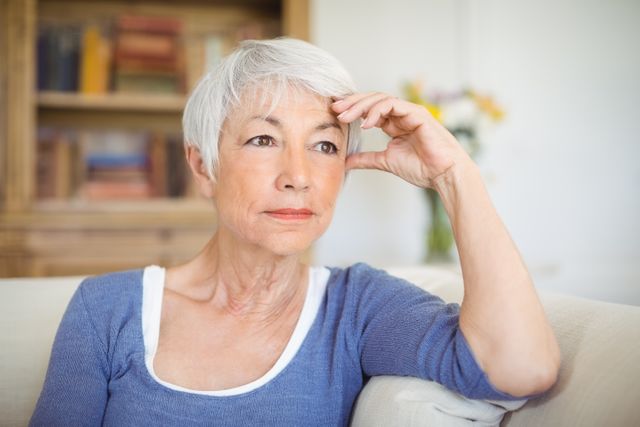 Senior woman with grey hair sitting on a sofa in a living room, appearing thoughtful and pensive. Ideal for use in articles or advertisements related to aging, retirement, mental health, lifestyle, and home living. Can also be used in healthcare and wellness contexts to depict contemplation and peacefulness.